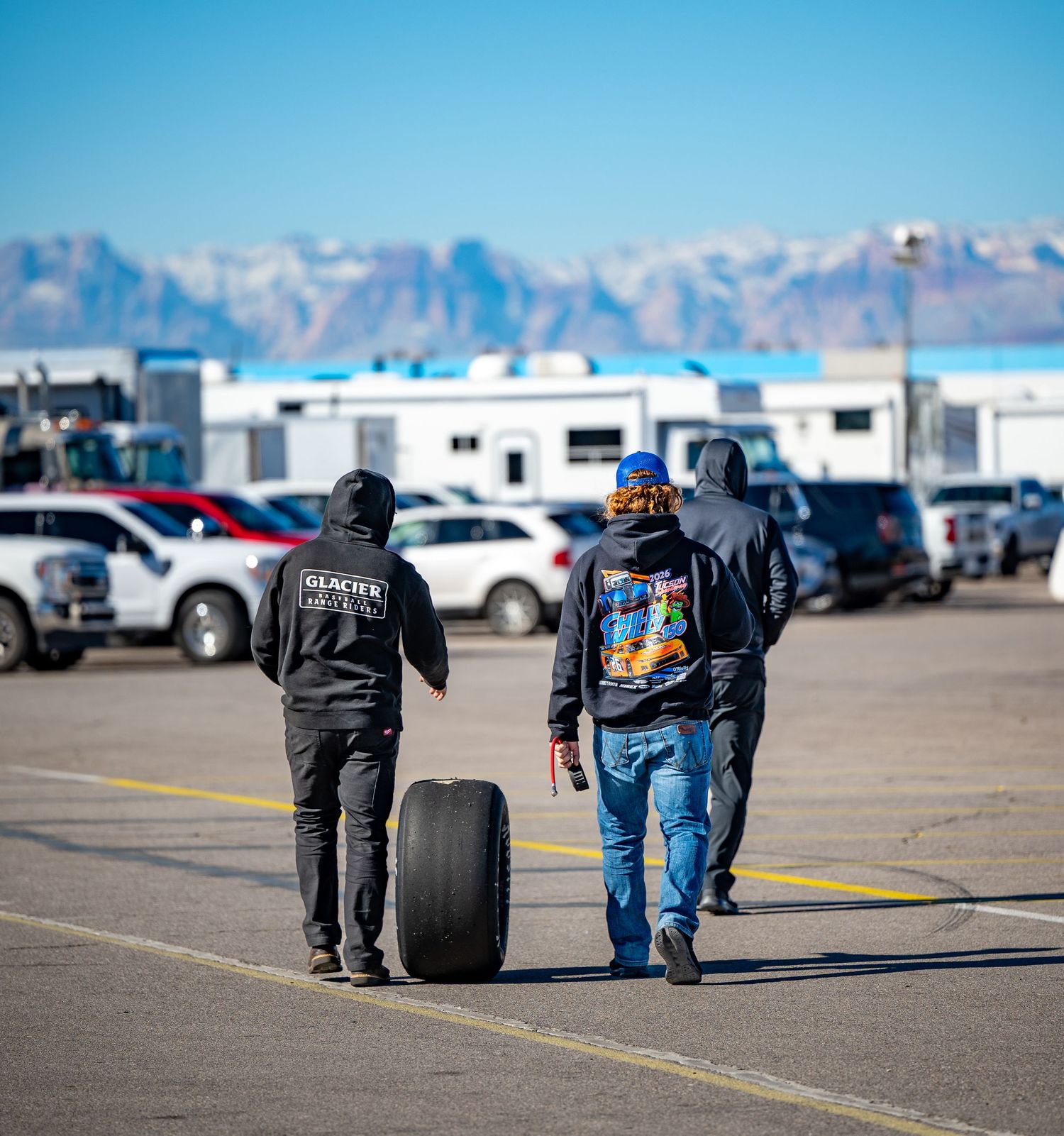 Two crew members roll a racing slick tire across paddock with mountain backdrop at motorsport event.