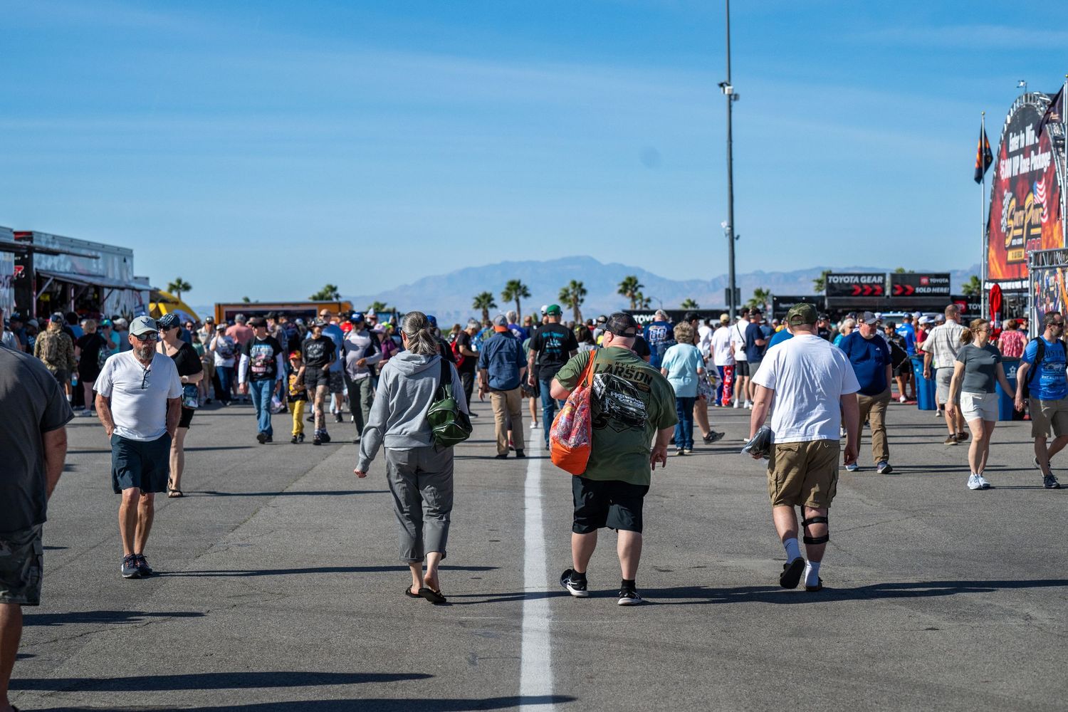 Large crowd of NASCAR fans walking across sun-drenched speedway infield with mountains in background.