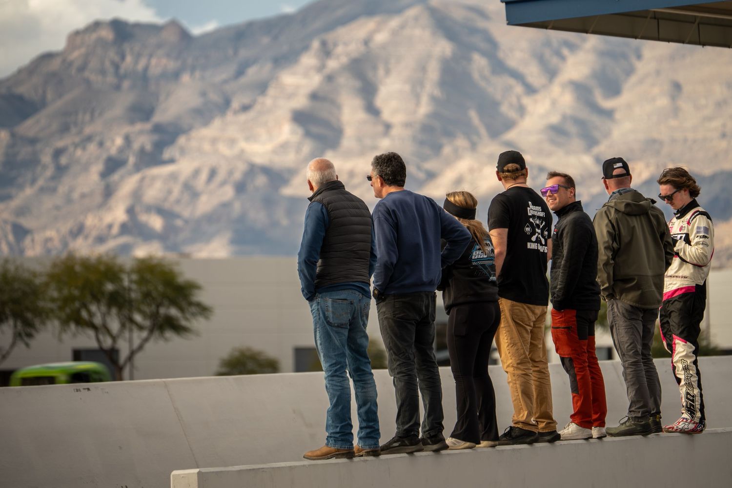 Group of spectators standing on a wall watching a racing event with mountain backdrop in desert setting.