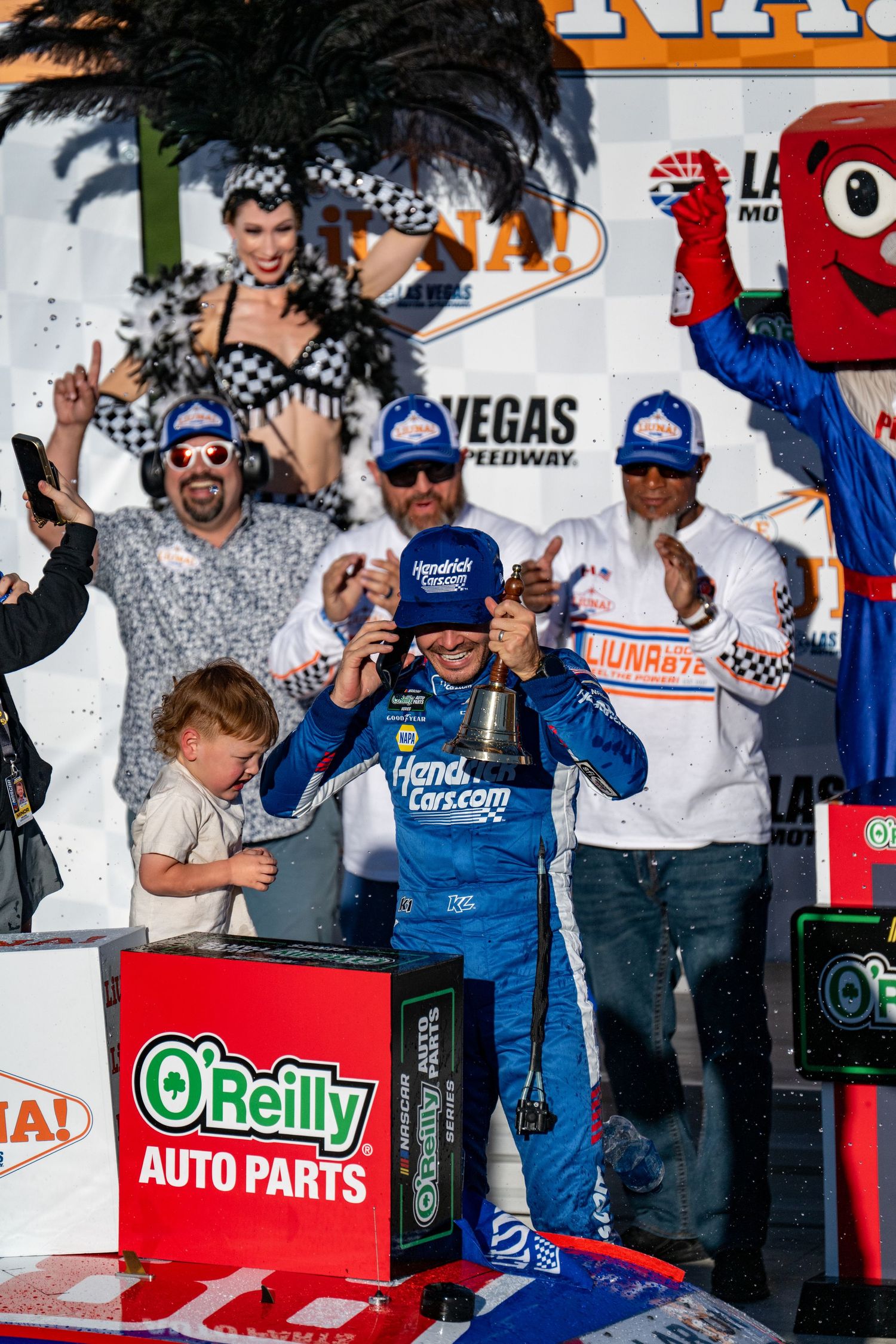 NASCAR driver celebrates victory in winner circle at Las Vegas Motor Speedway with trophy bell.