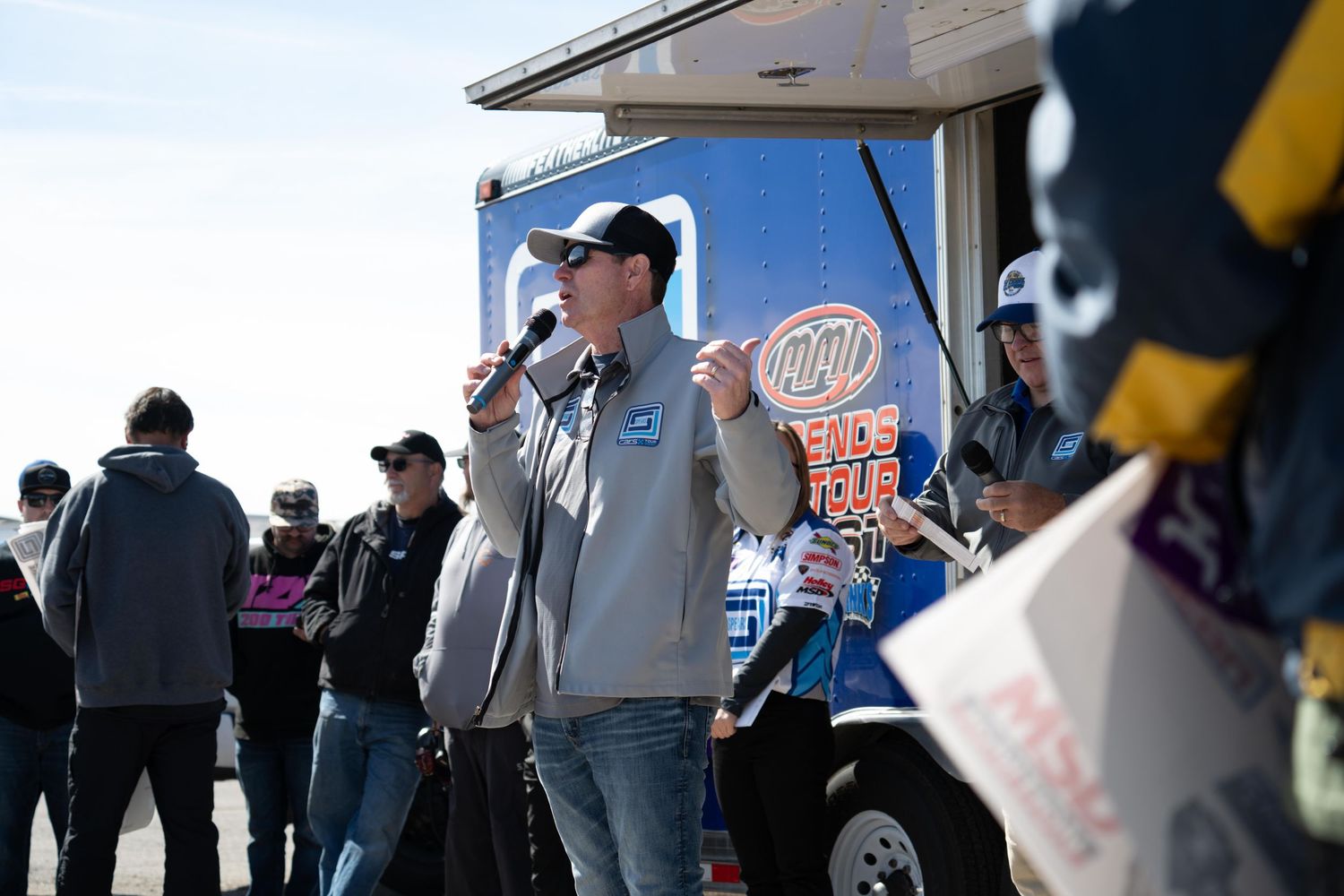 Man speaks into microphone at outdoor NASCAR fan event near branded promotional truck.