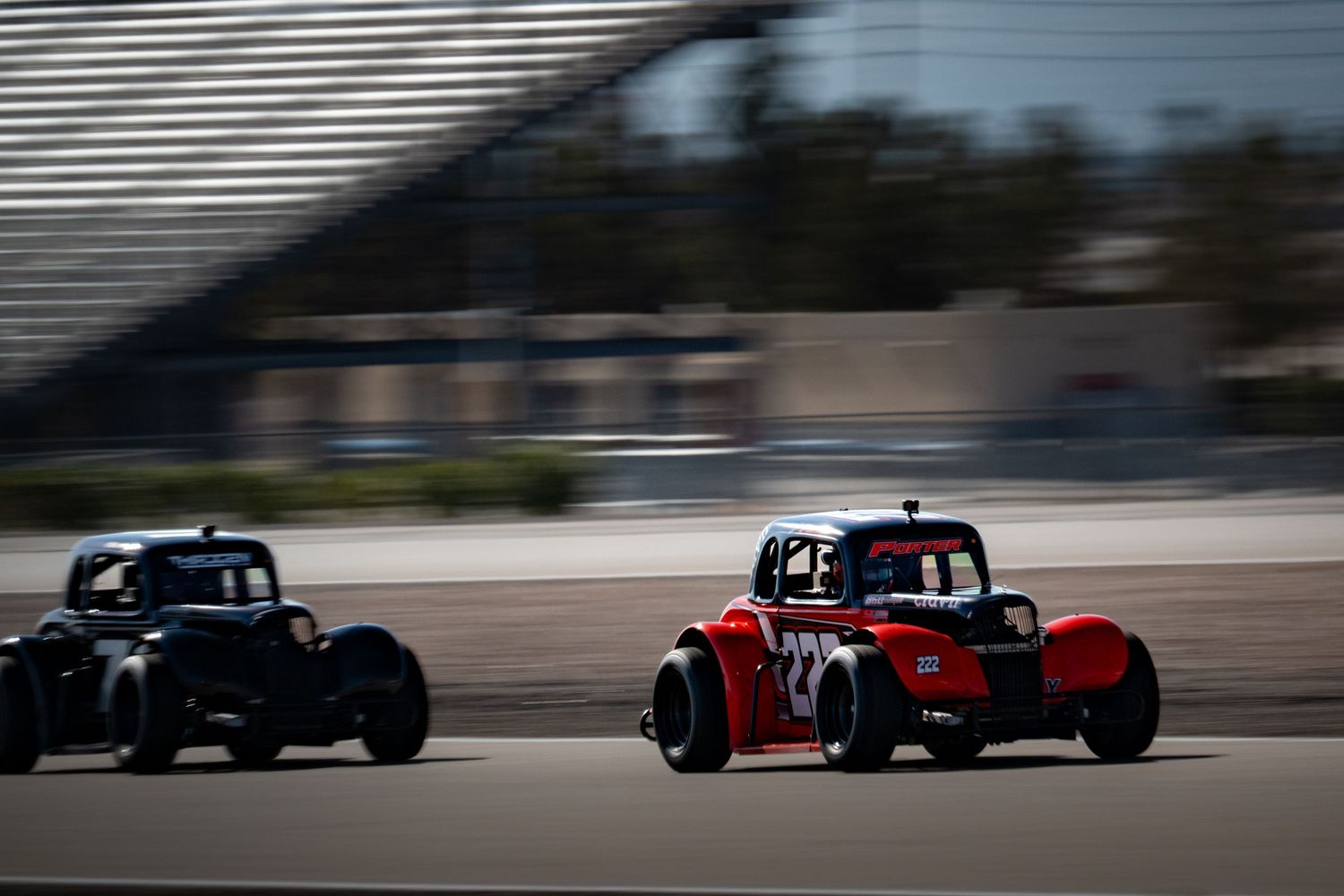 Two vintage-style legend cars racing on a wide oval track with grandstands visible.