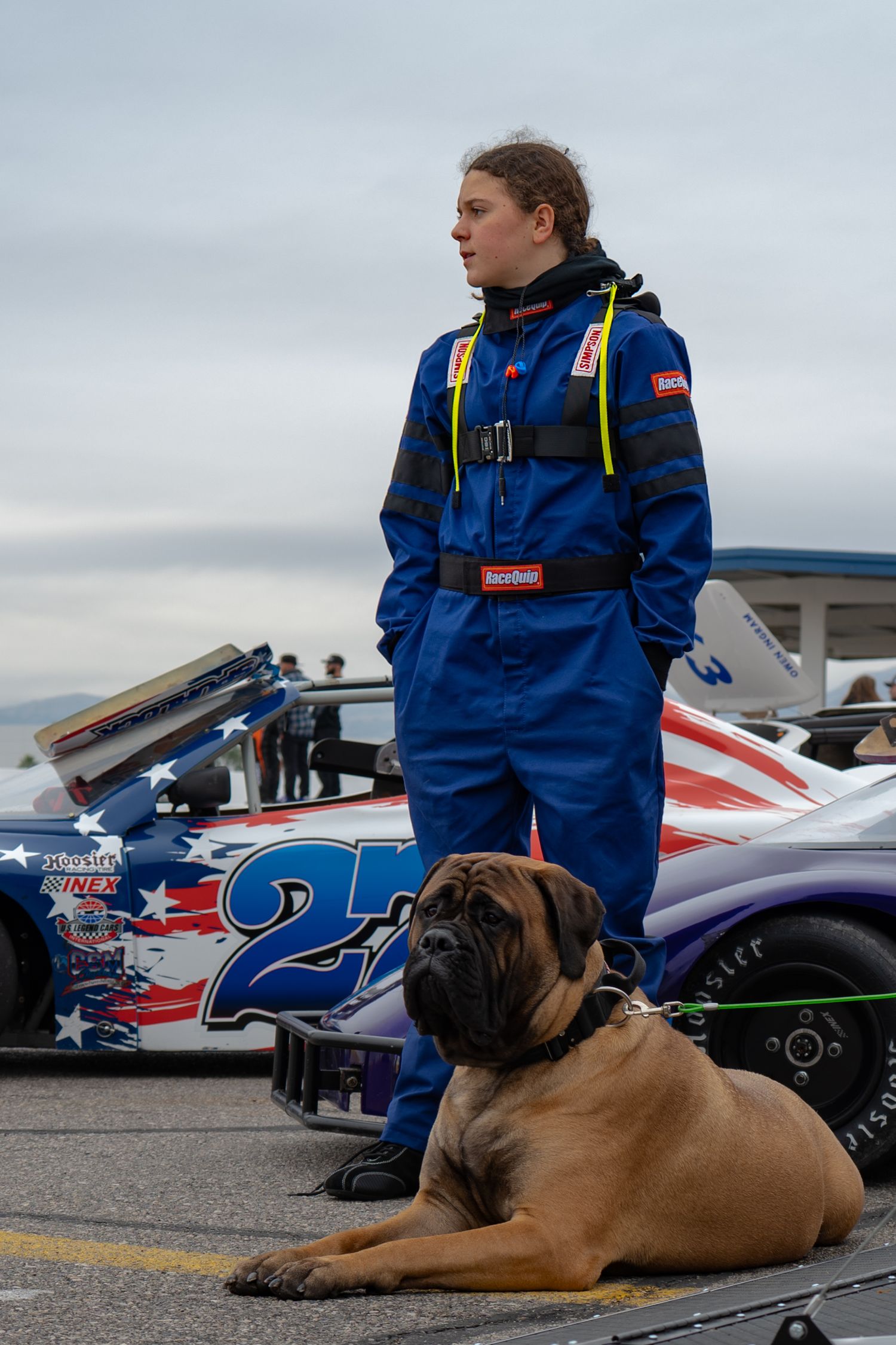 Female race car driver in blue firesuit standing beside car number 27 with large mastiff dog.
