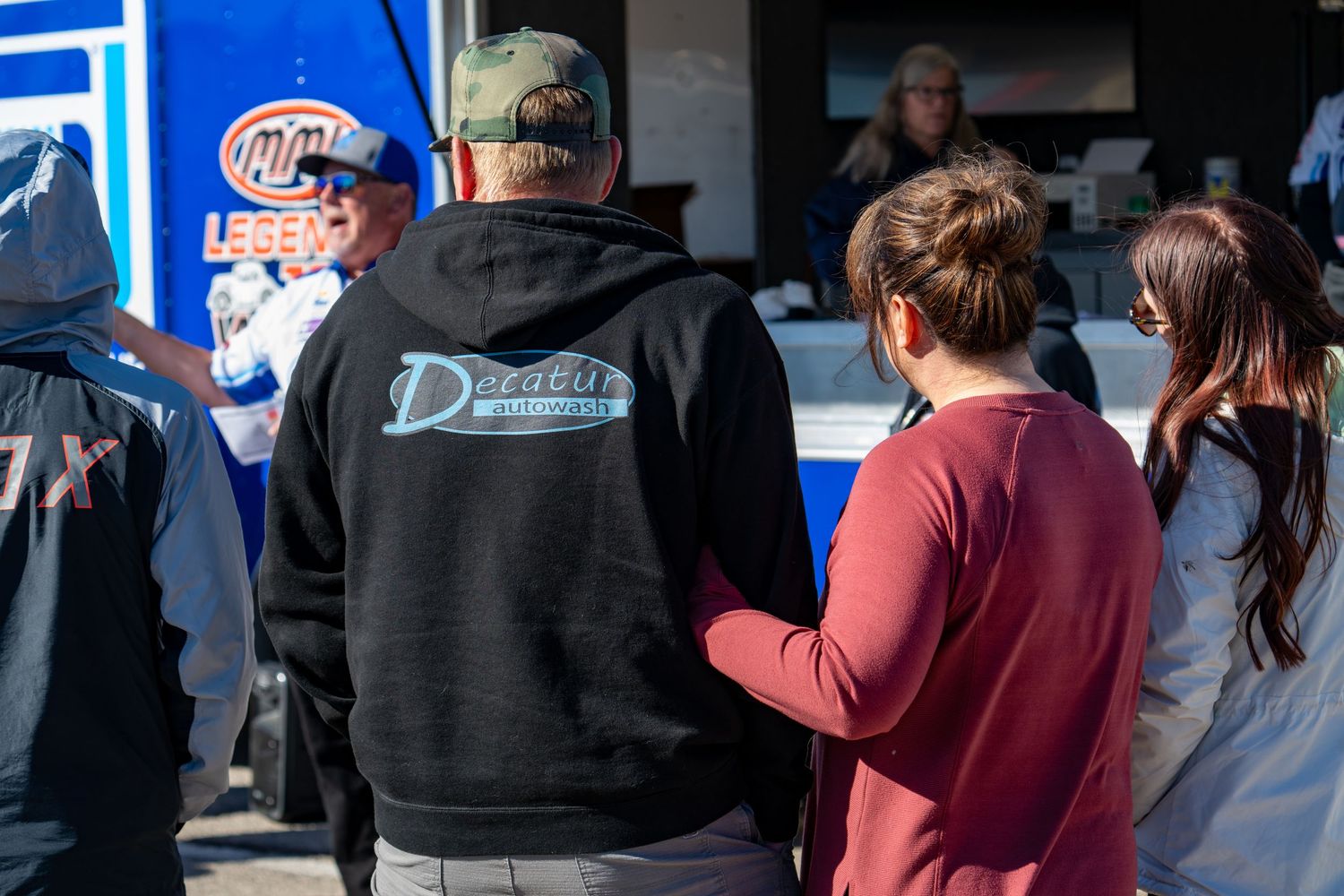 People wearing racing team hoodies gather at an outdoor motorsport event near a sponsor booth.