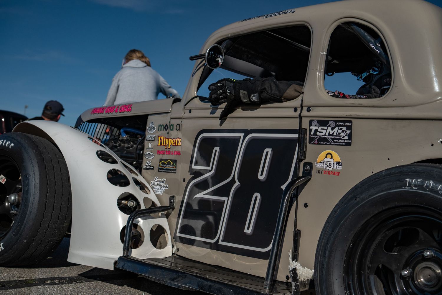 Close-up of a vintage-style gray racing car numbered 28 with sponsor decals at a motorsport event.