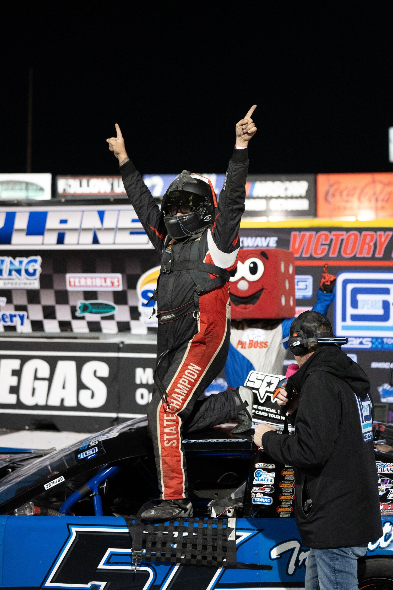 Race driver celebrates victory standing on car with both arms raised in Las Vegas victory lane.