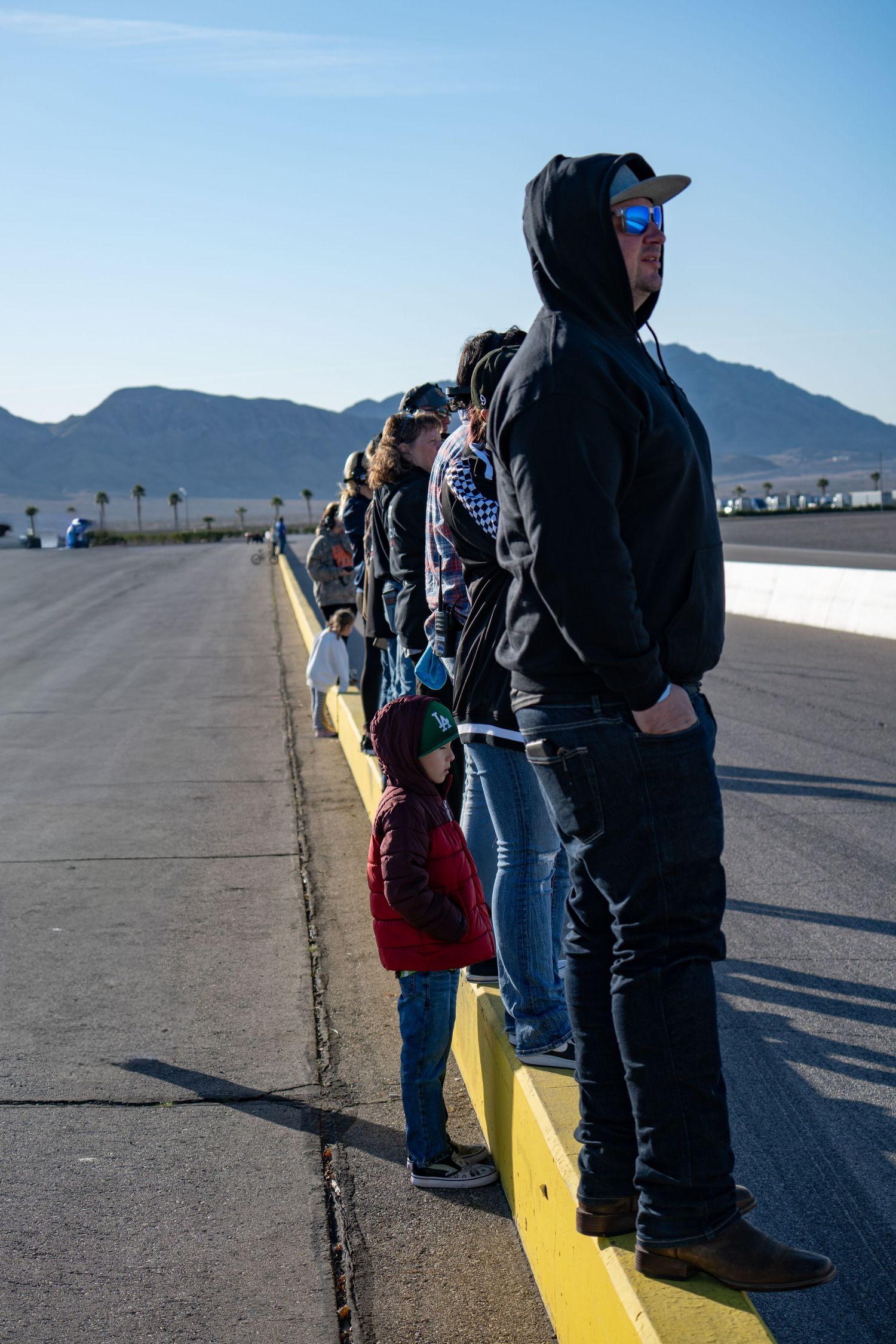 Group of spectators including a child standing along a yellow pit lane barrier at a desert racetrack.
