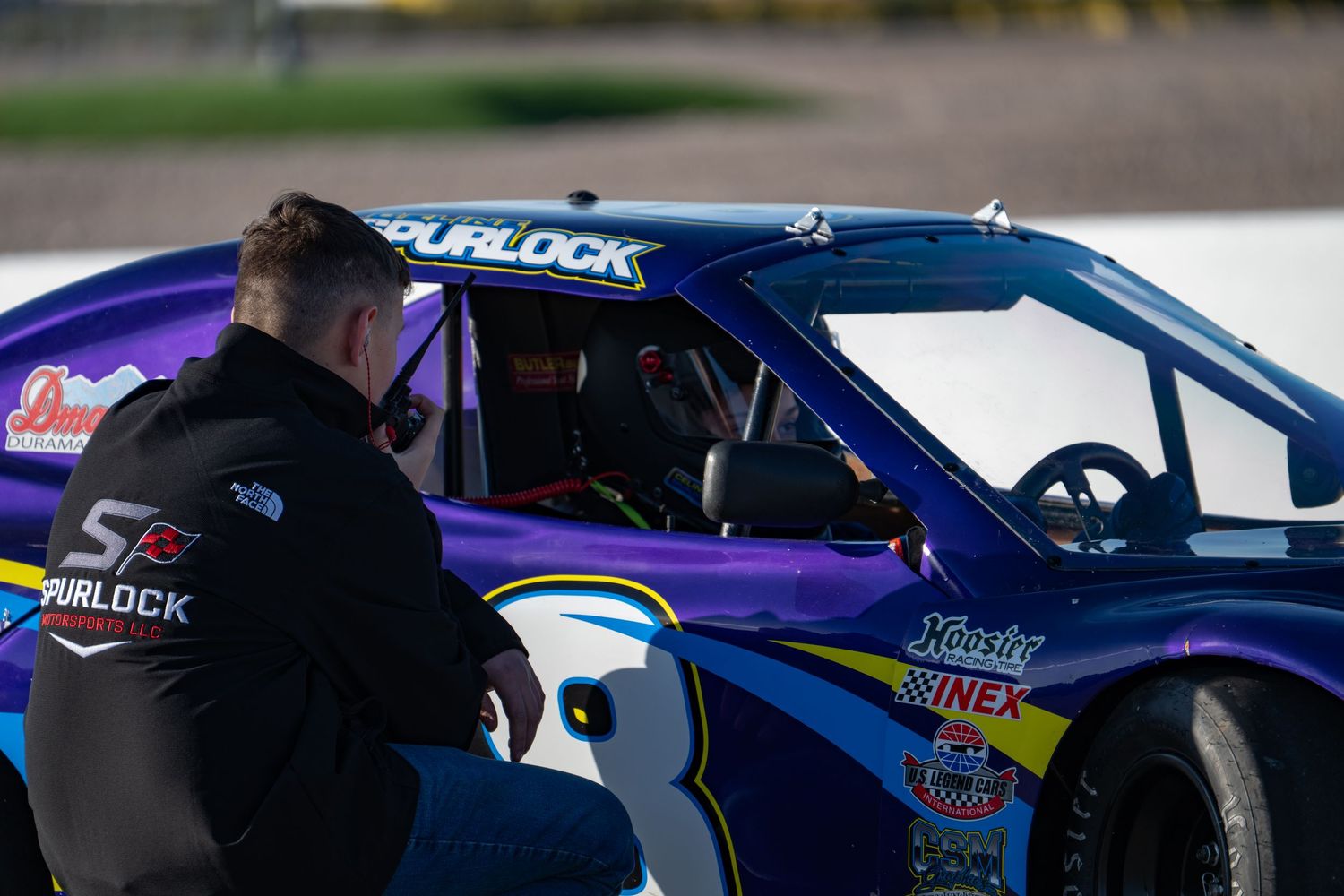 Racing crew member in Spurlock jacket crouching beside blue Legend Car number 8 on pit lane.