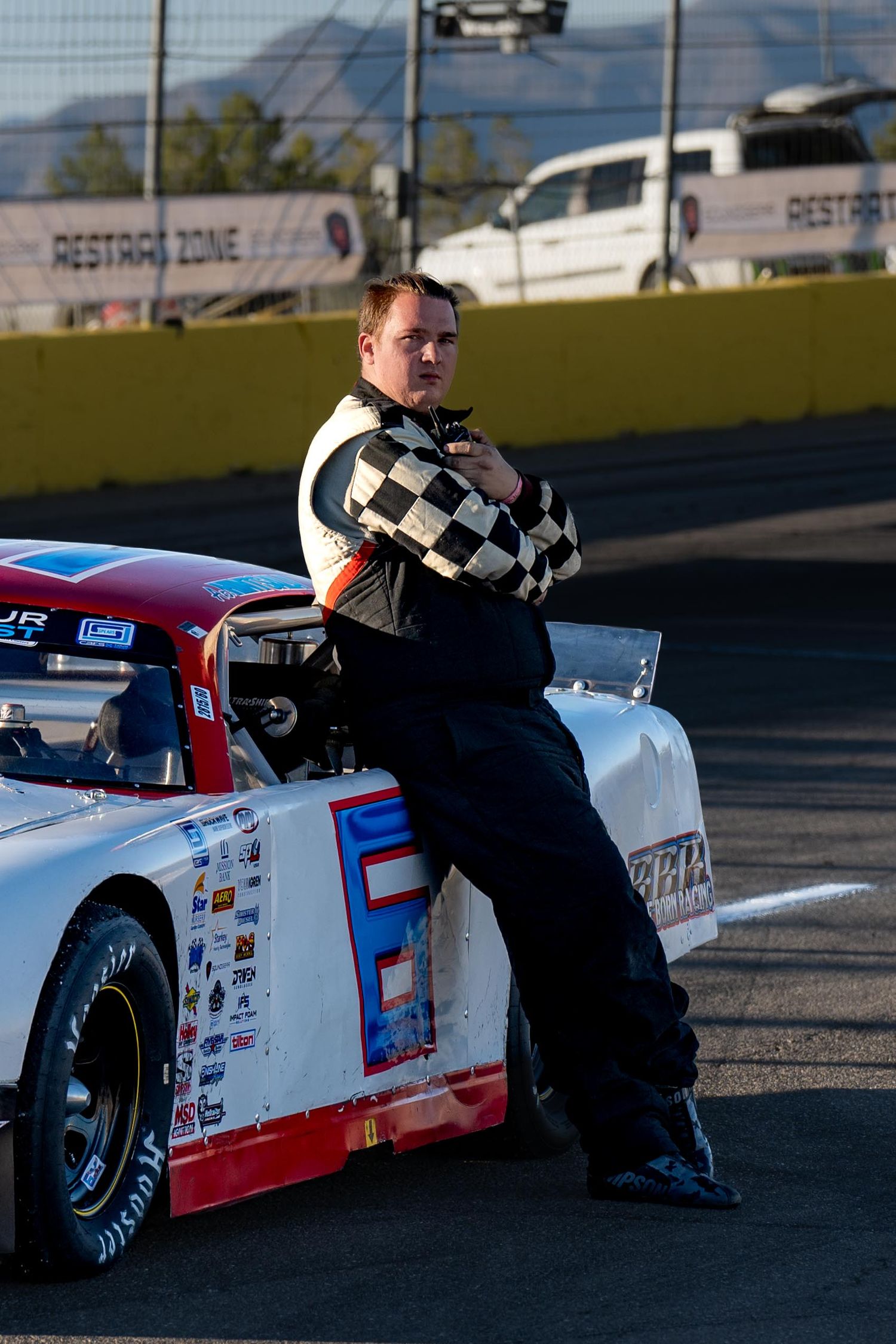 Male race driver in checkered flag racing suit leans against red and white stock car on track.