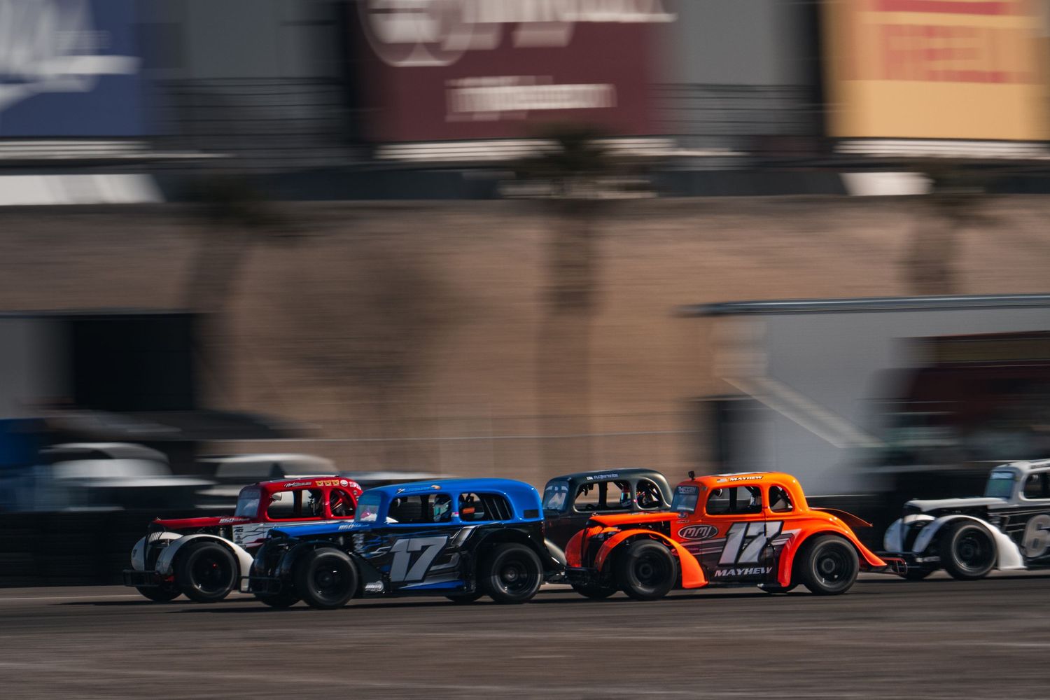 Three vintage-style legend cars racing side by side on an oval track at dusk.