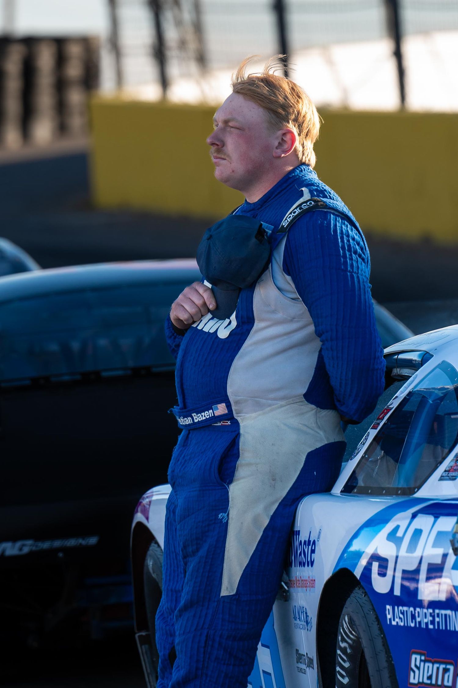 Race car driver in blue firesuit leaning against car at Las Vegas Motor Speedway during sunset.