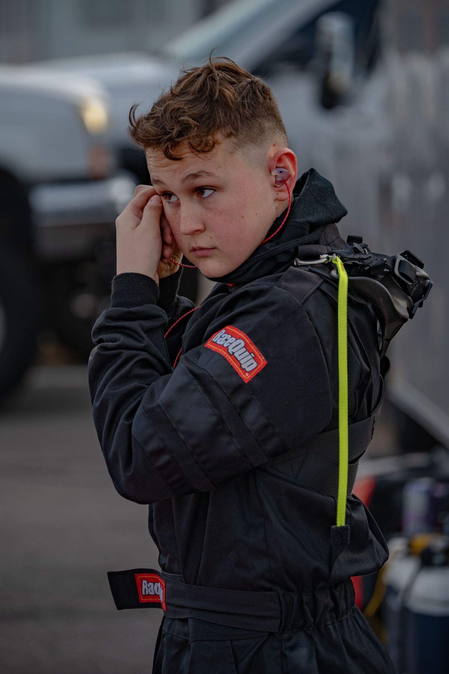 Young boy in RaceQuip racing suit touching his ear while standing in a race paddock area.