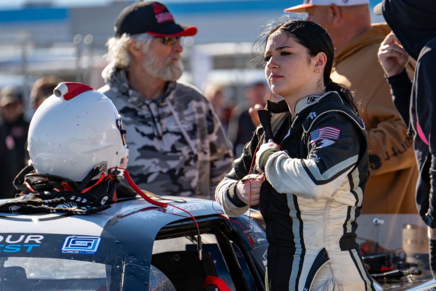 Female race driver in black racing suit stands beside car at motorsport event with crew nearby.