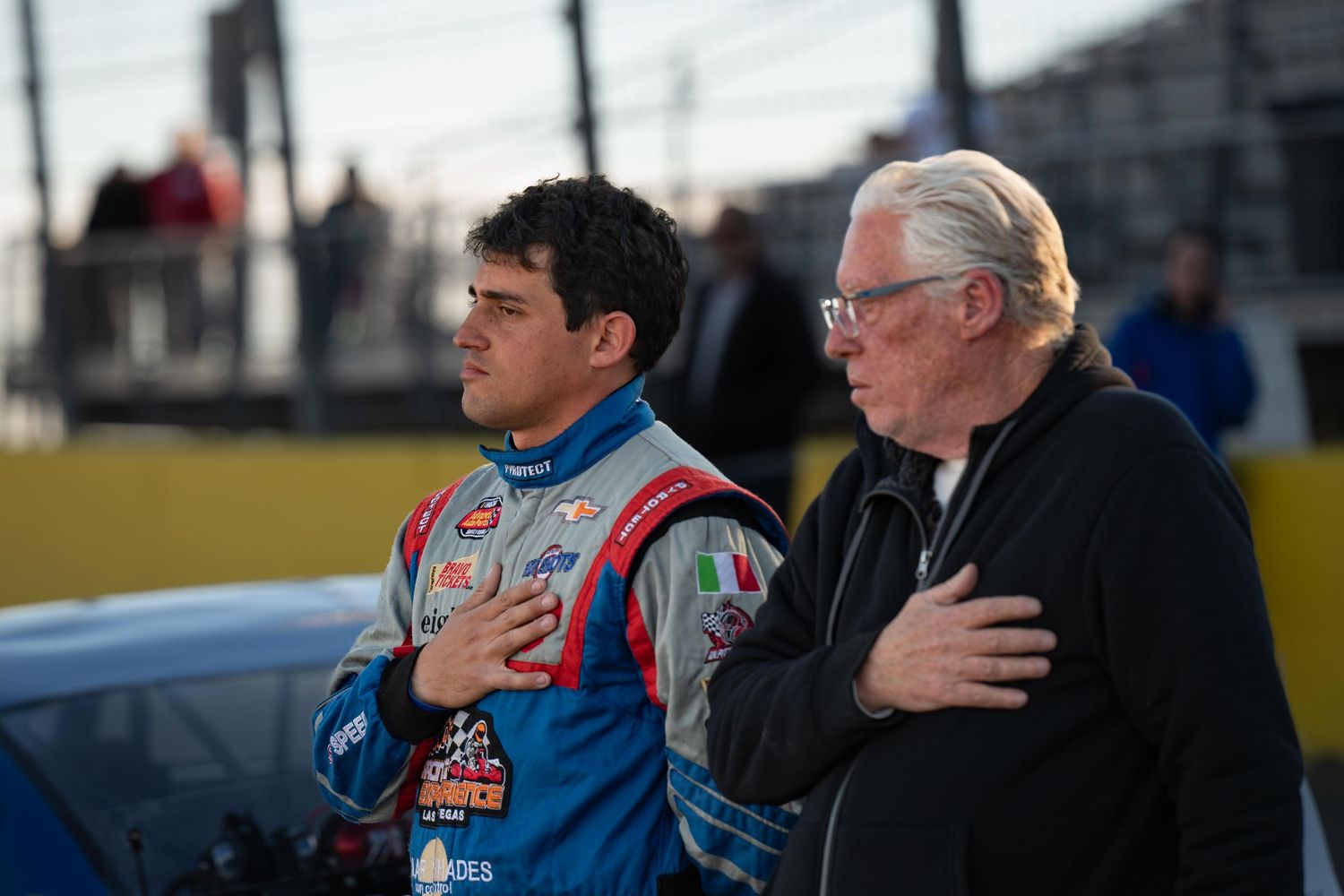 Father and son stand with hands over hearts during national anthem at motorsport event.
