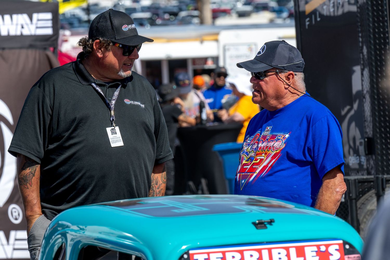 Two men in caps have a conversation beside a turquoise classic car at NASCAR vendor booth for US Legends and INEX