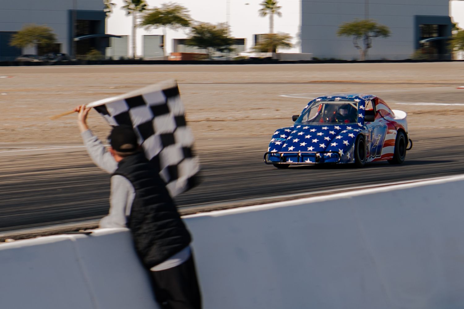 Race official waves checkered flag as patriotic American flag bandolero car crosses finish line at sunset.