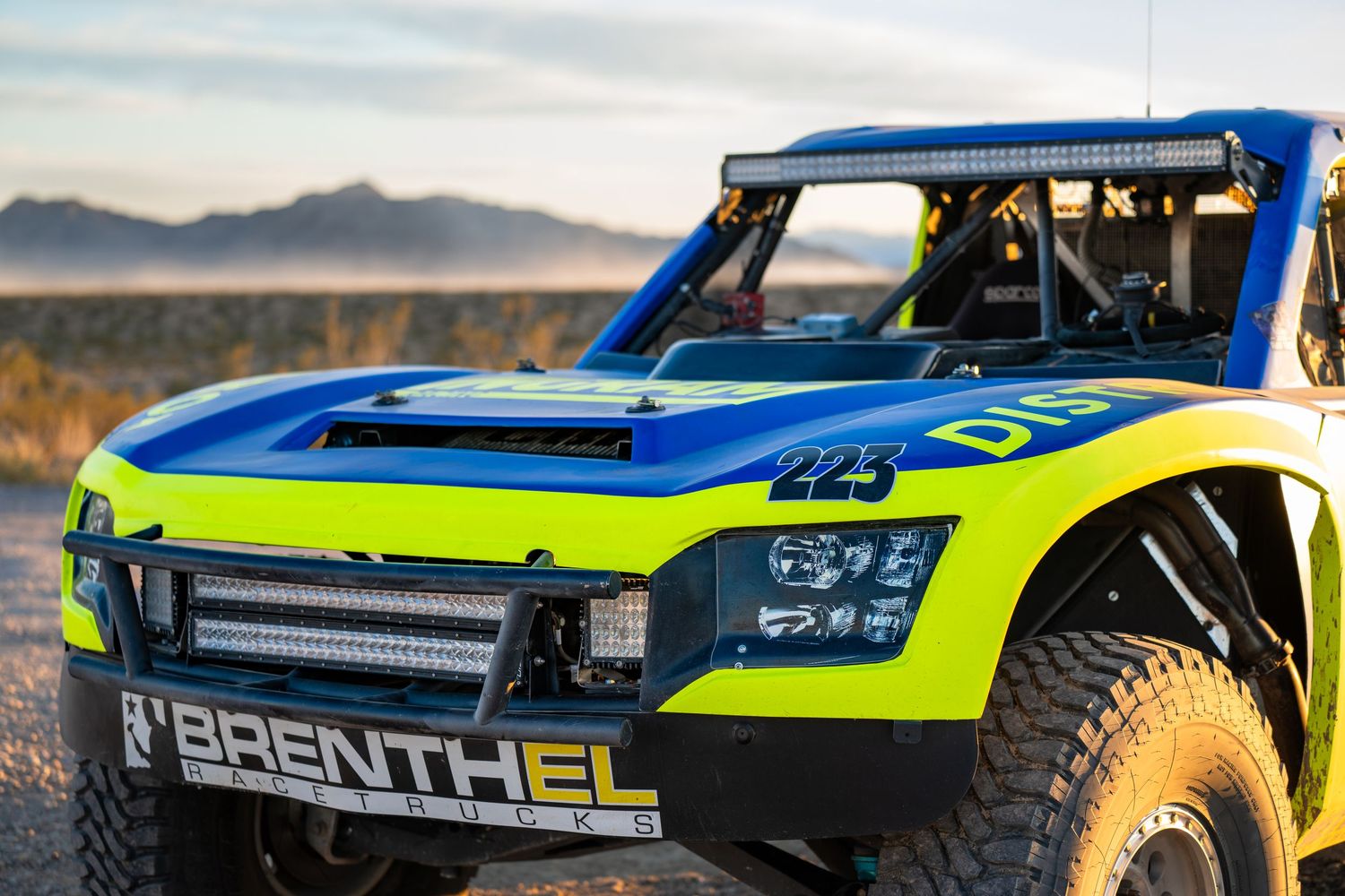 Front detail of blue and green Brenthel trophy truck number 223 with LED light bar in desert.