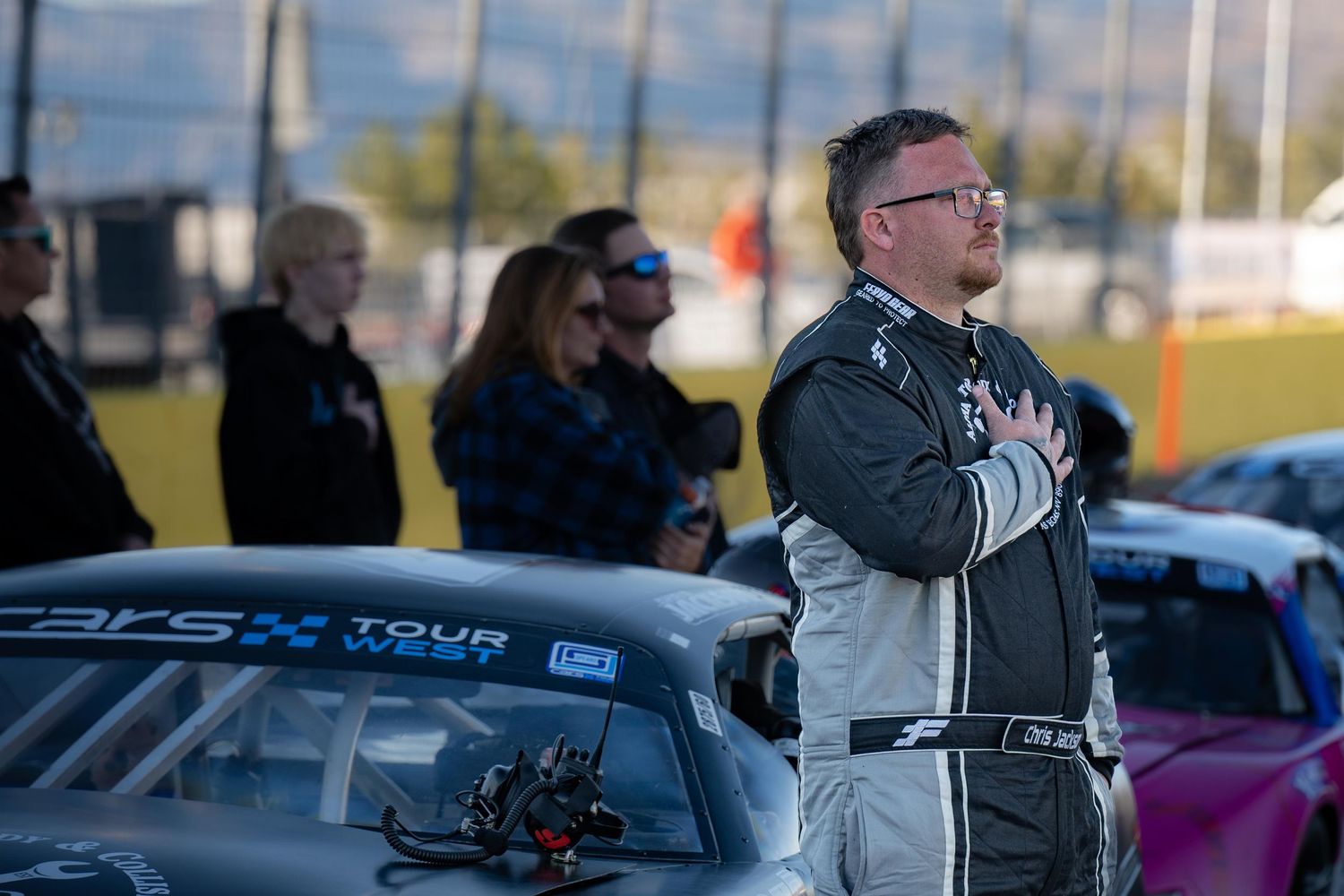 Male race driver in gray suit stands with hand on chest during national anthem at racing event.