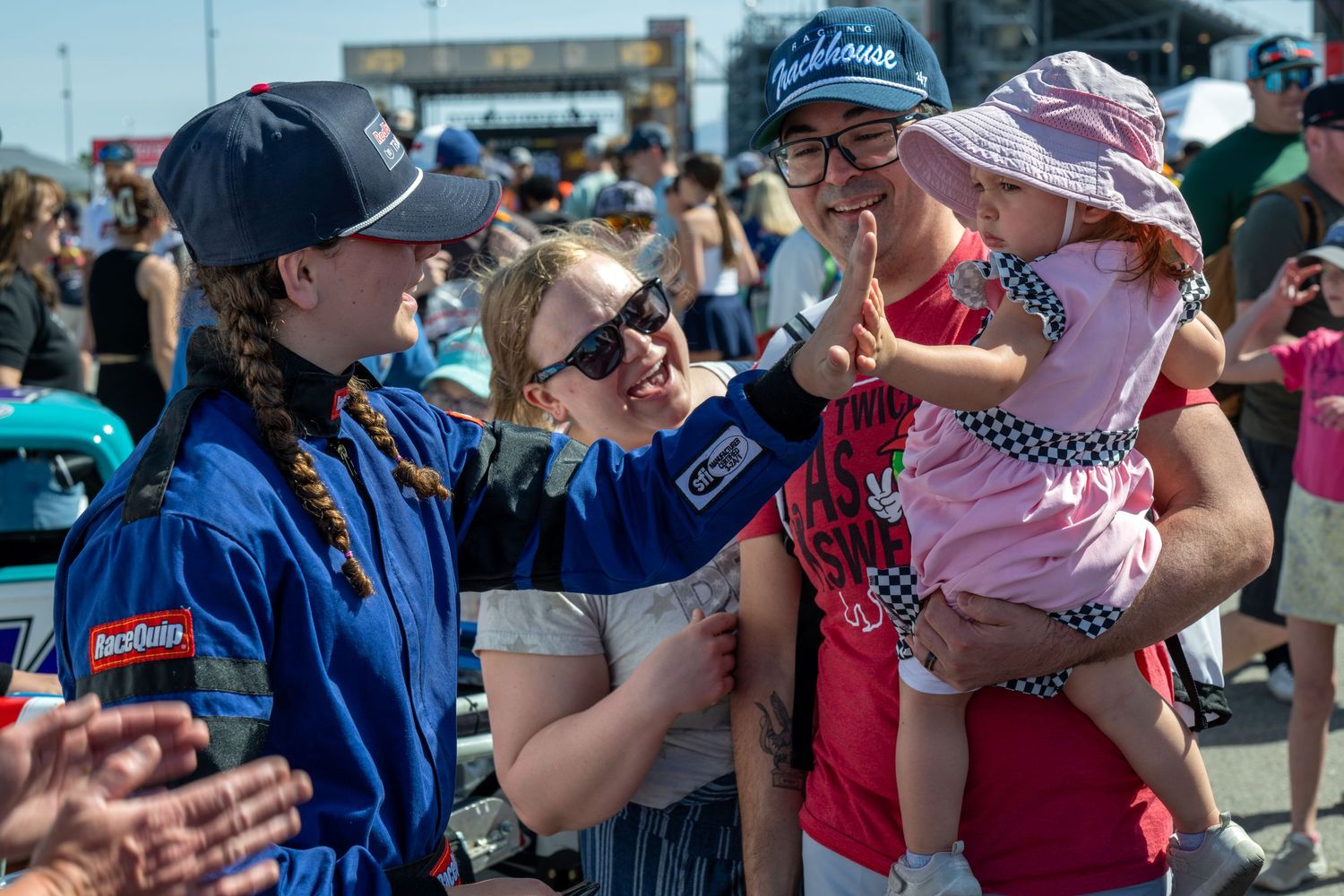 NASCAR driver in blue racing suit greeting fans including woman holding baby at speedway event.