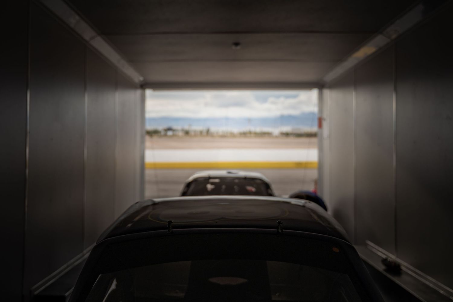 Dark interior view of a race car loaded inside a silver enclosed trailer with track visible outside.