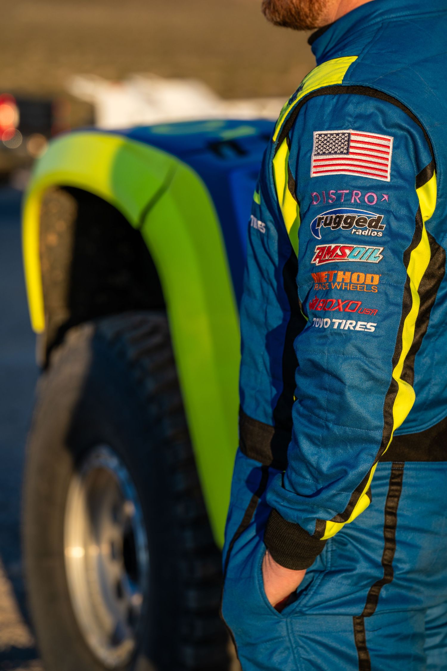 Close-up of a racing driver's blue suit sleeve showing sponsor logos including Distro and Rugged Radios.
