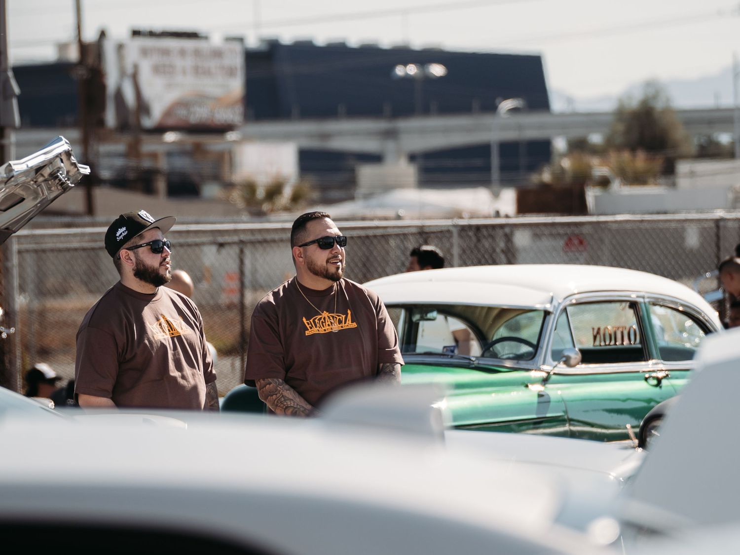 Two men in matching brown shirts standing near vintage classic cars at Super Saturday Weekend at The STRAT in Las Vegas