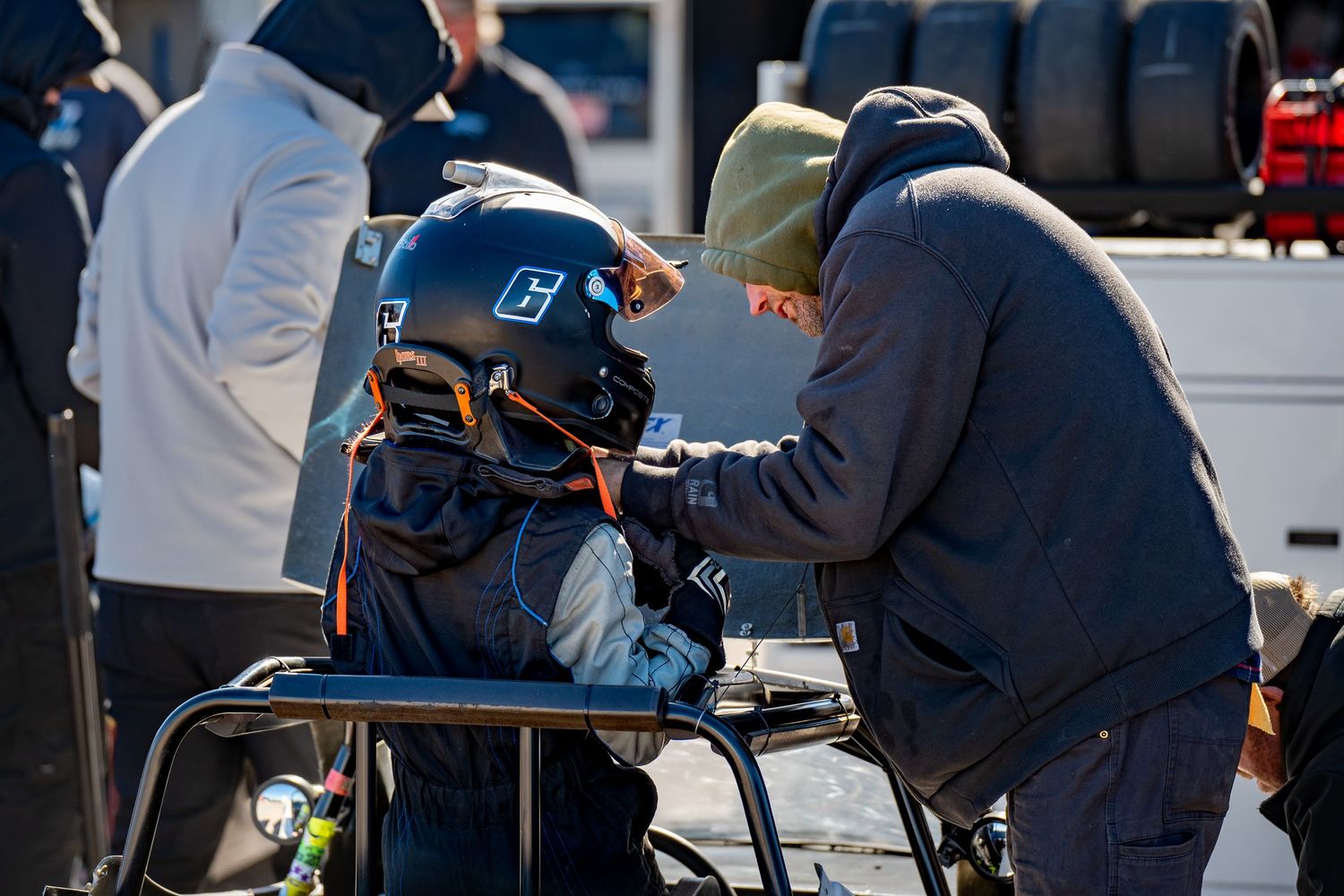 Racing crew member adjusts helmet of young driver seated in go-kart at motorsport event.