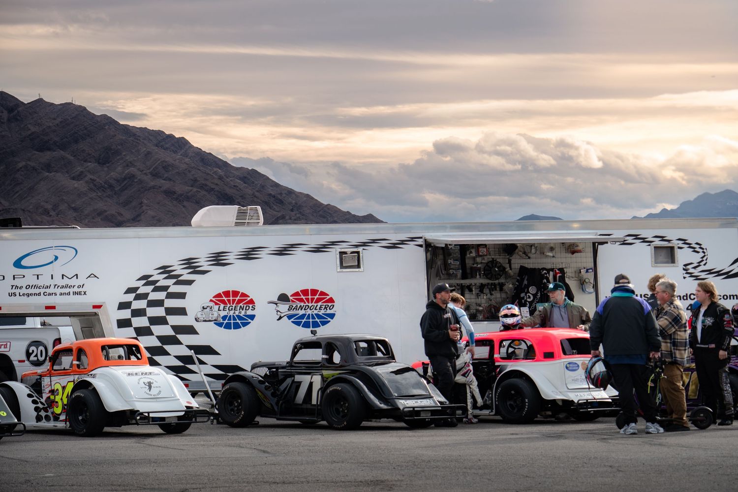 Vintage legend race cars parked near a racing trailer at dusk with mountains and lake in background.