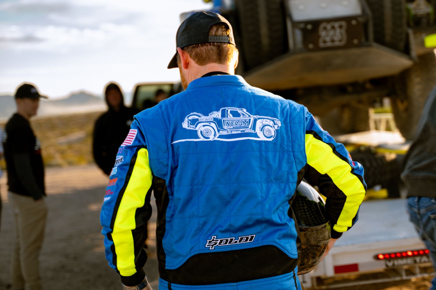 Rear view of a racing driver in a blue and yellow Ingram Racing suit with a truck graphic at a desert race event.