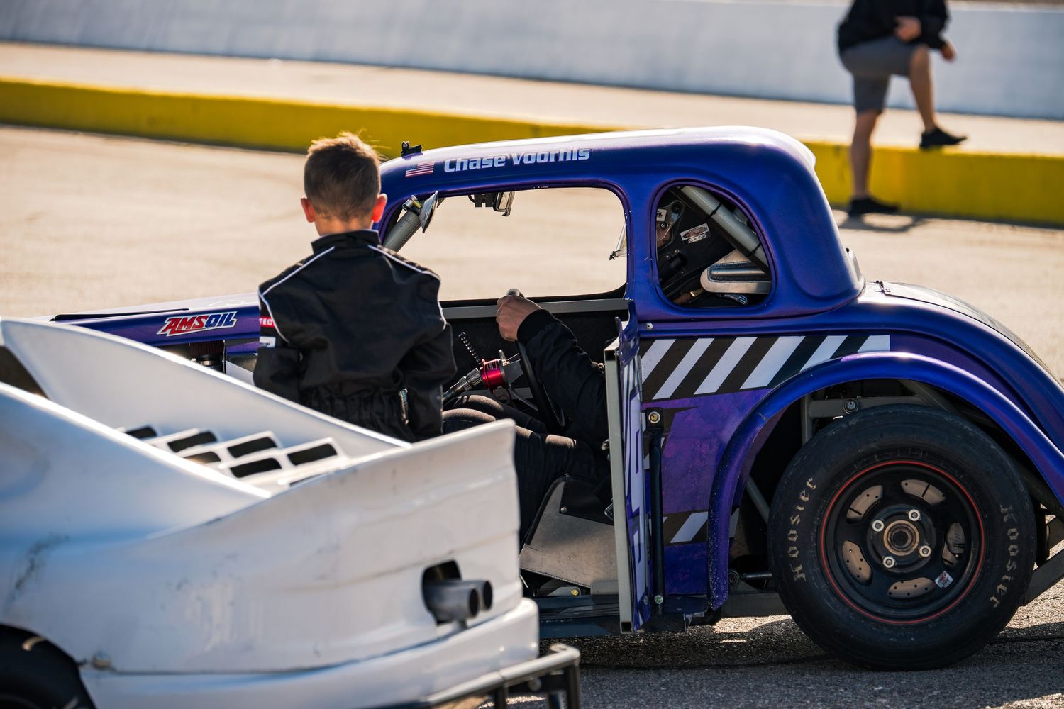 Young child driver sitting in a small blue Legend Car in the racing paddock area.