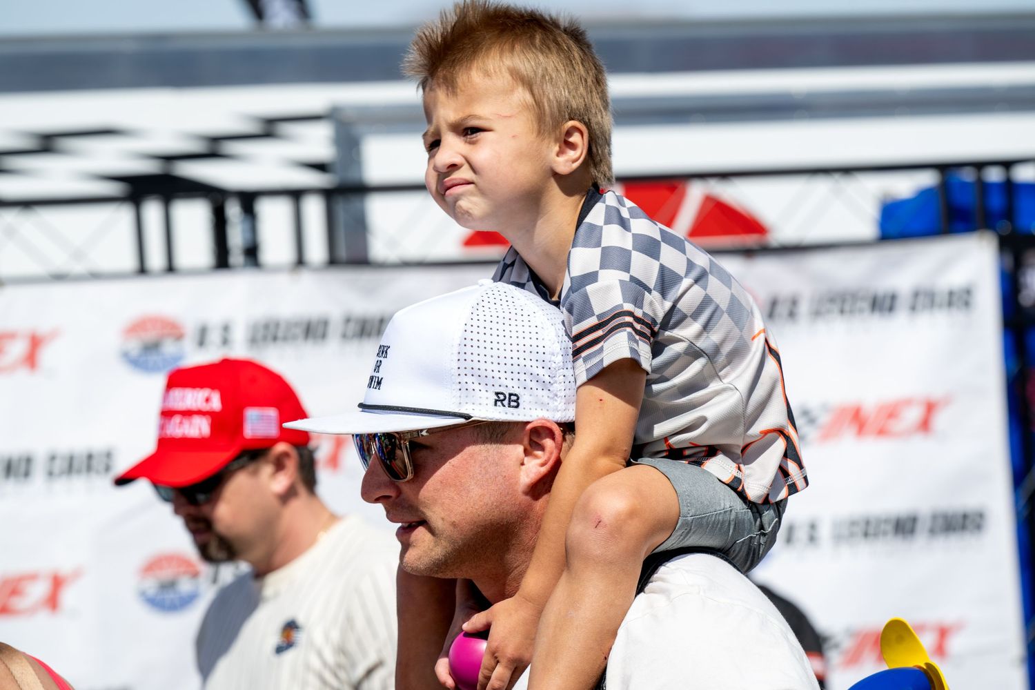Young boy sits on an adult's shoulders wearing a checkered shirt at NASCAR event