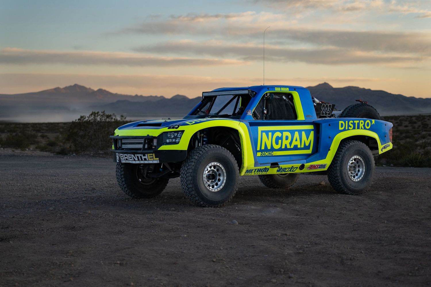 Blue and yellow Ingram Distro off-road trophy truck parked in desert at dusk with mountain backdrop.
