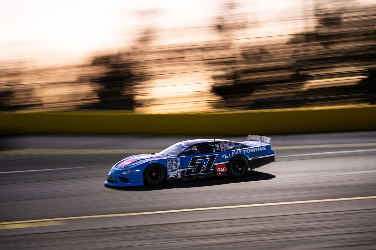 Blue number 57 stock car racing at sunset on an oval track with motion blur capturing high speed.