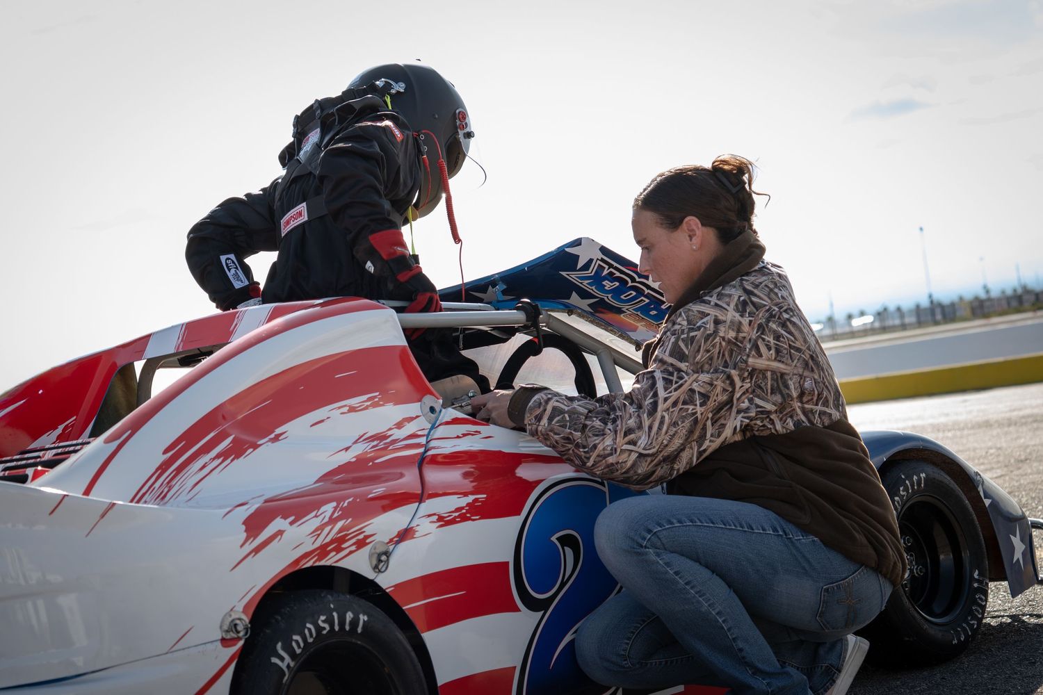 Woman crouching beside white number 2 junior race car giving instructions to a helmeted young driver.