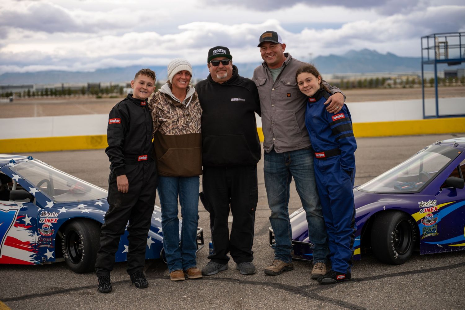 Group of five people posing together between two race cars on a racing track.