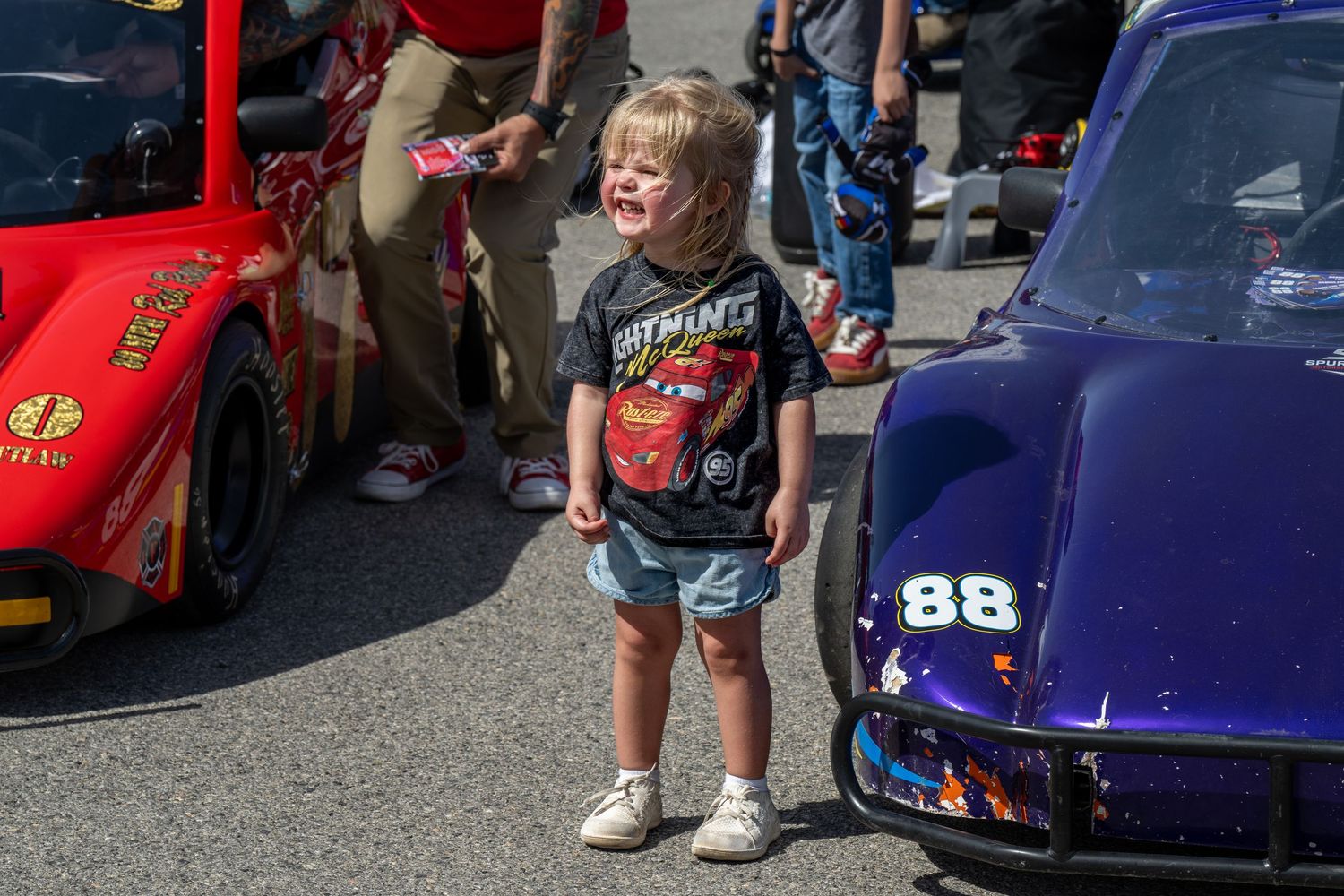 Excited toddler in Lightning McQueen shirt stands between colorful race cars numbered 88 at a racing event.