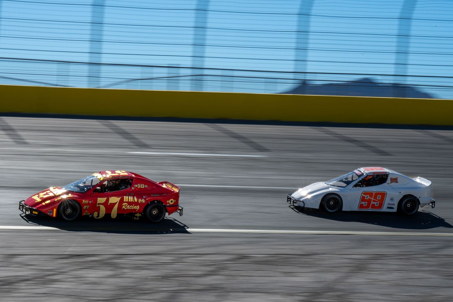 Red number 57 and white number 99 race cars competing side by side on Las Vegas bullring oval short track.