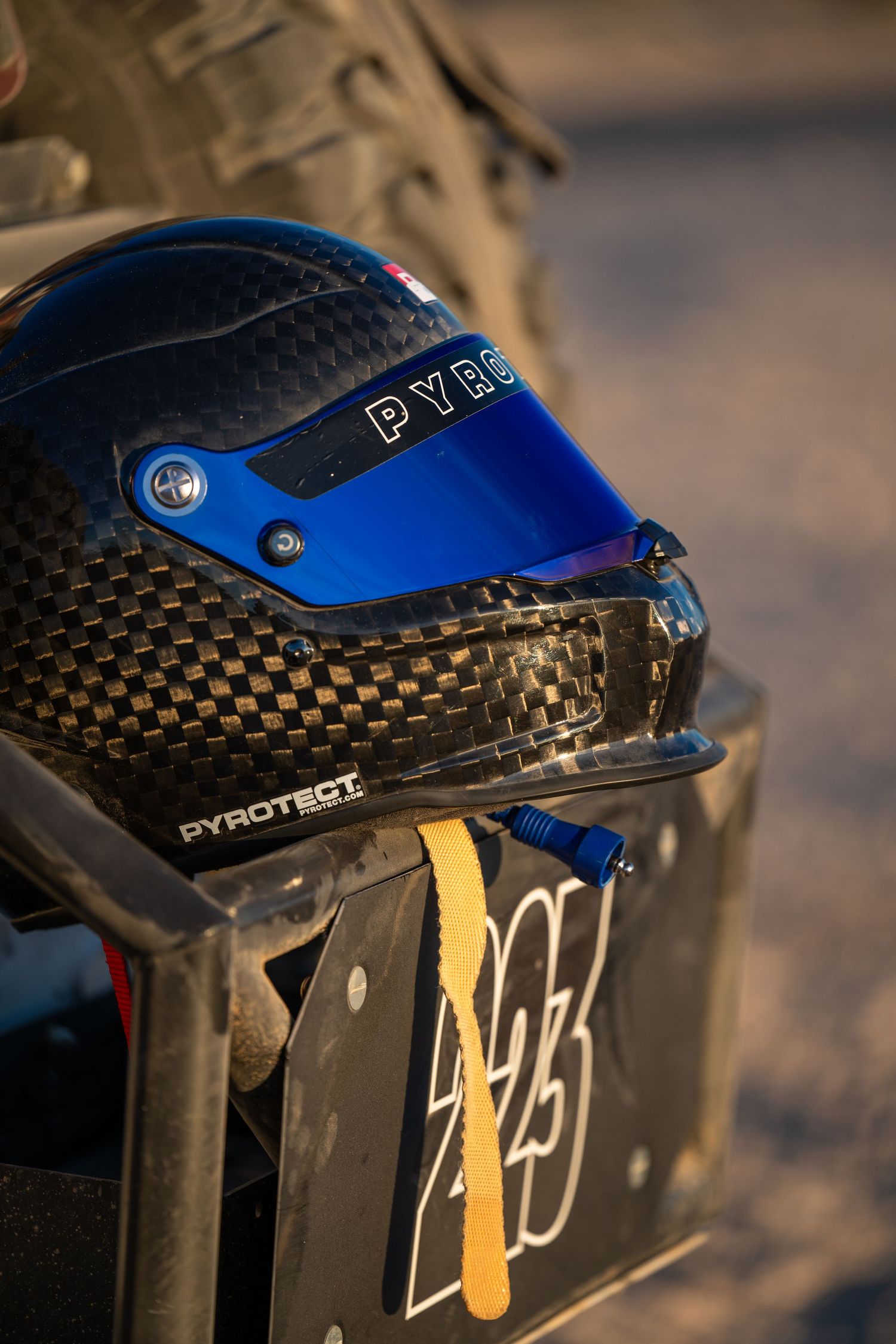 Pyrotect carbon fiber racing helmet with blue visor resting on a trophy truck bumper.