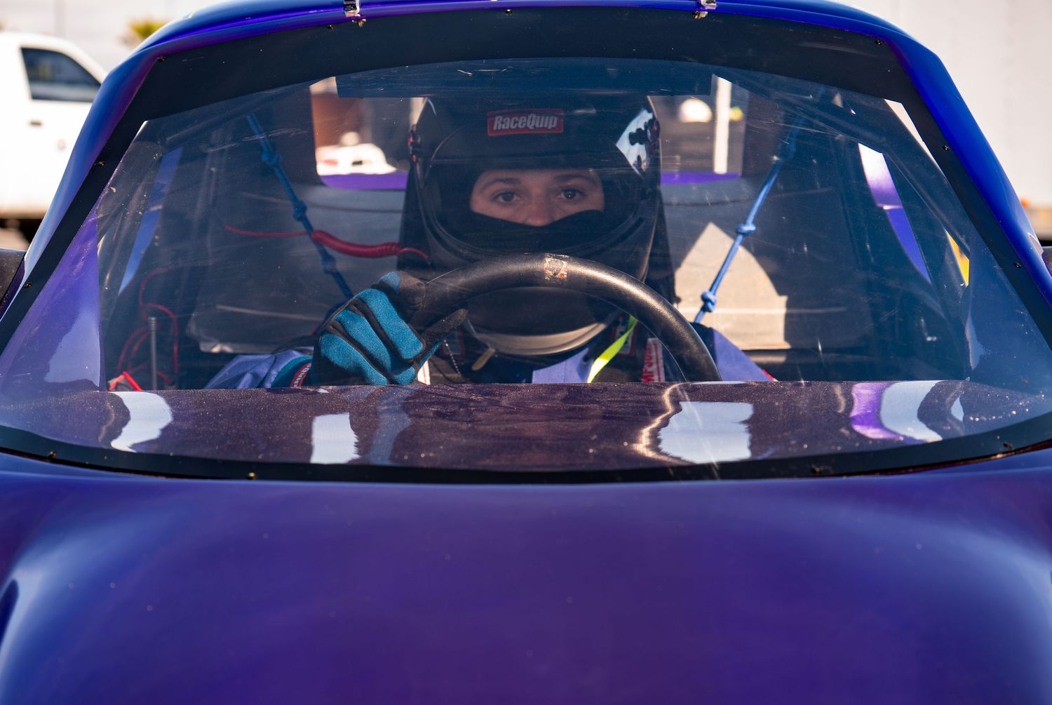 Female bandolero race car driver in helmet and gear sits ready behind steering wheel of purple stock car.