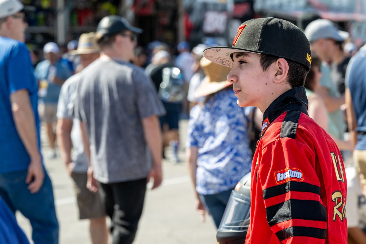 Young male NASCAR driver in red racing jacket and black cap at busy speedway fan event.