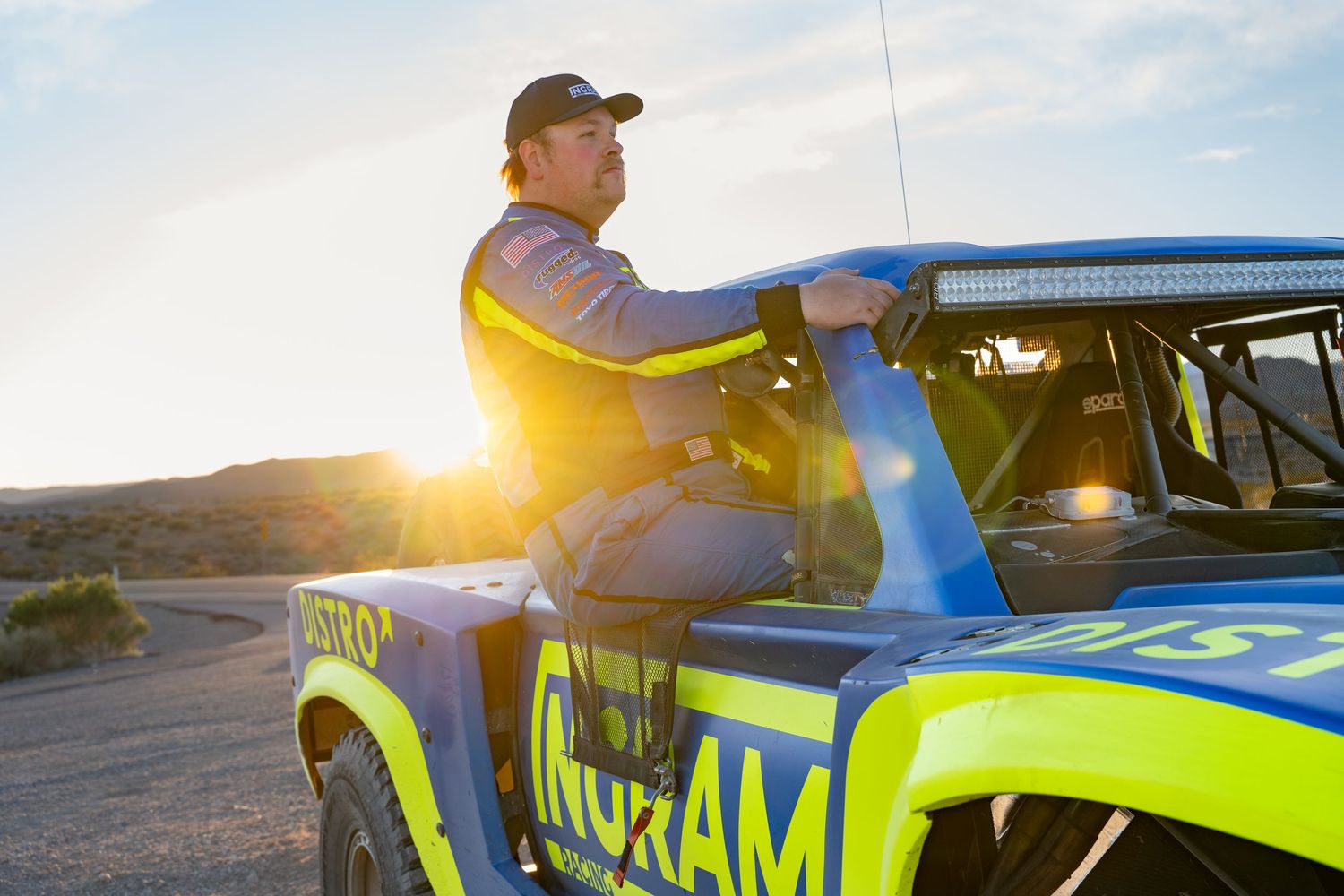 Racing driver leaning on a blue and green Distro off-road trophy truck at golden sunset.