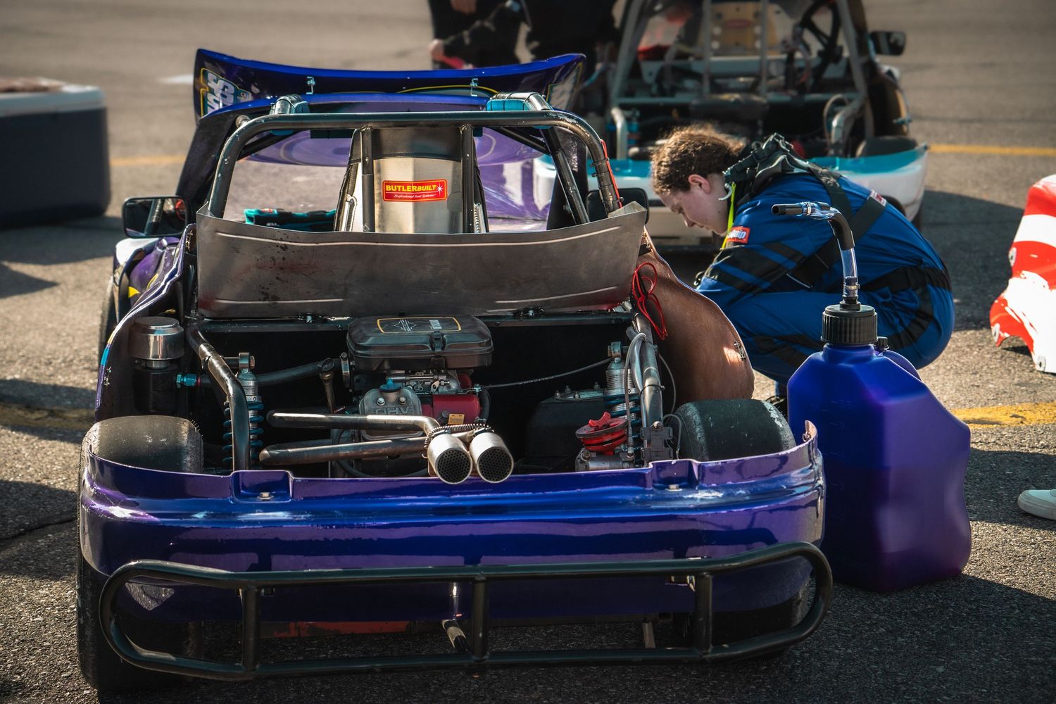 bandolero driver in blue racing suit works on engine of purple car with fuel can nearby on track.