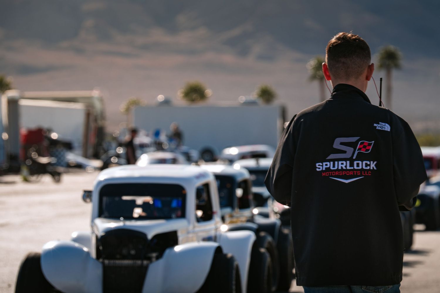 Man in Spurlock Racing jacket watching vintage-style race cars line up at a desert racetrack.