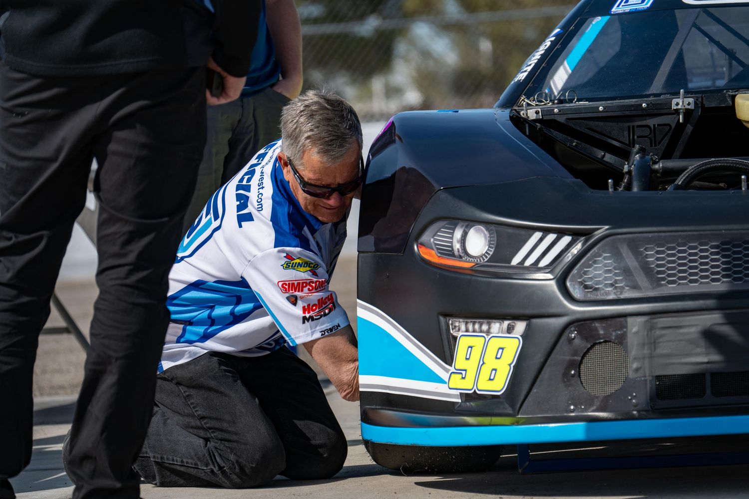 NASCAR crew member inspects number 98 race car during pit lane inspection at motorsport event.