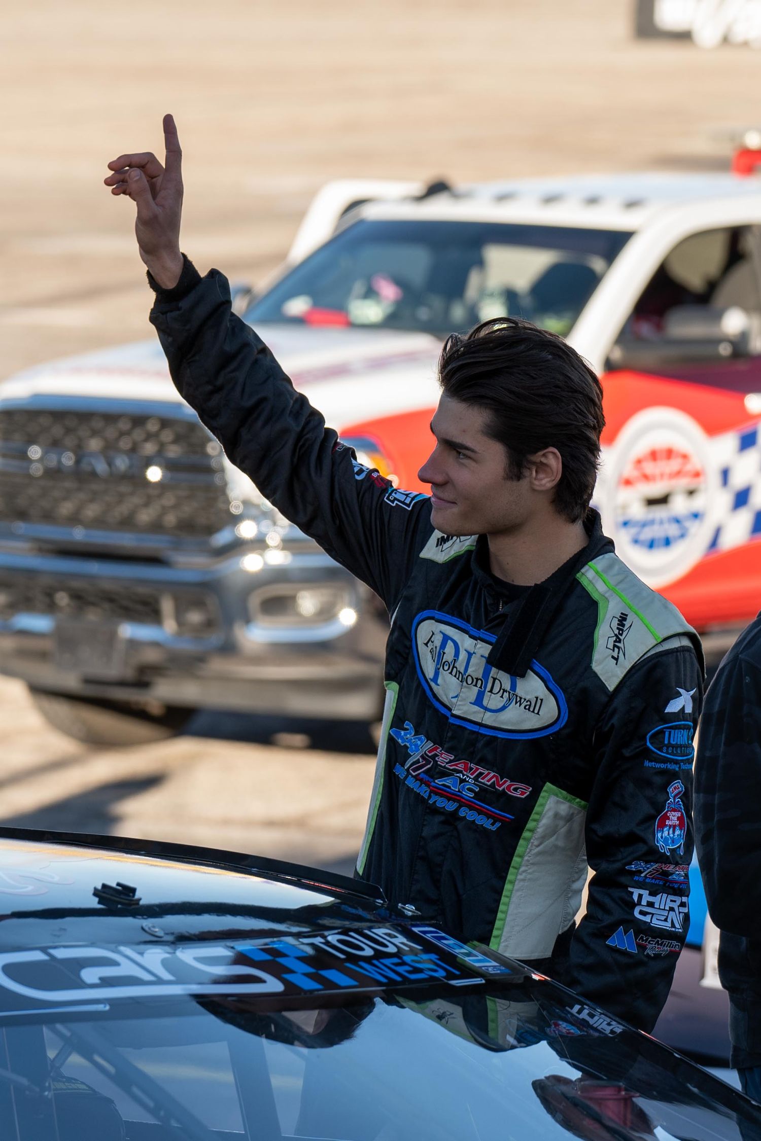 Young race car driver in black firesuit waving to crowd at motorsport event.