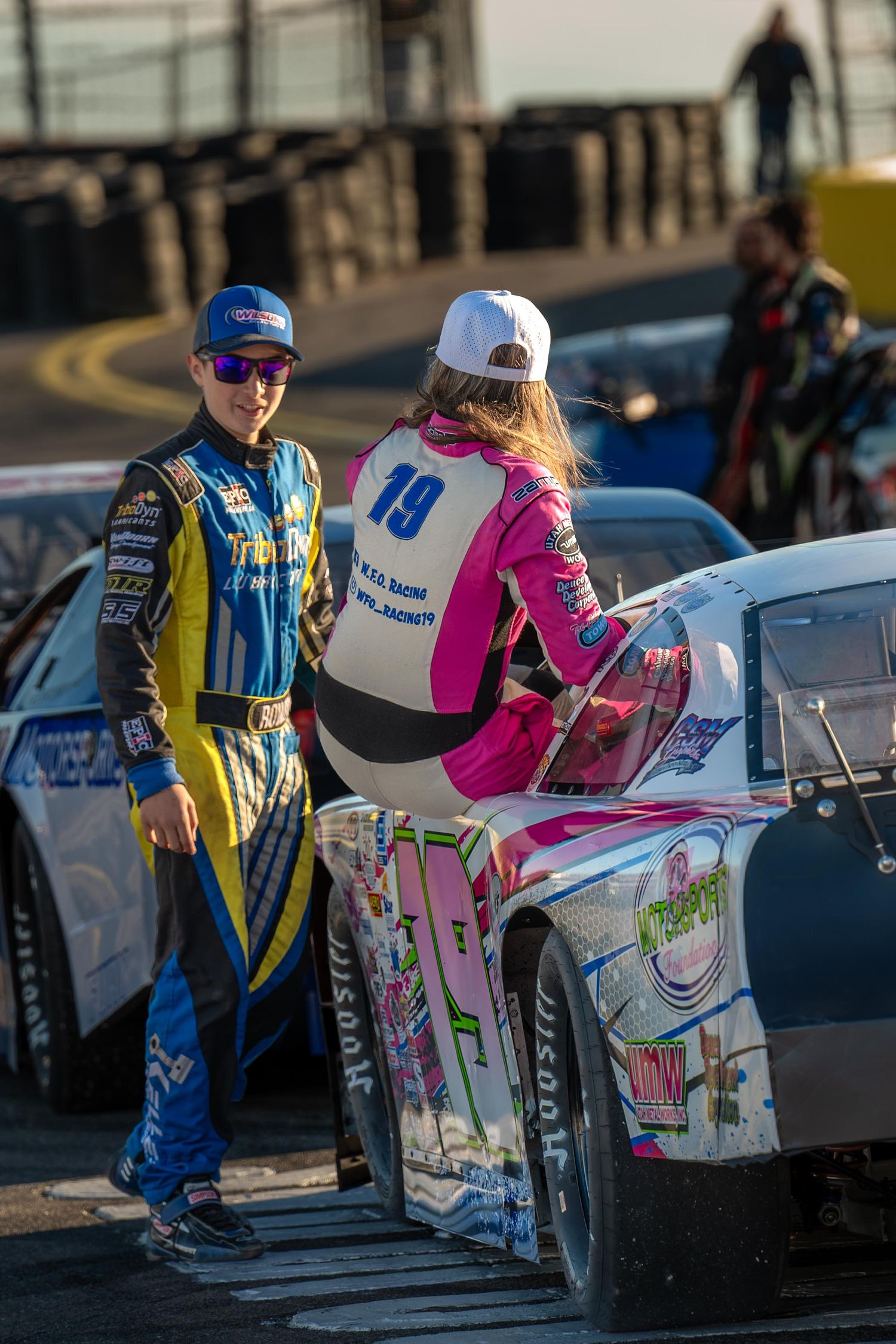 Two young race drivers in colorful racing suits converse beside number 19 stock car at sunset.