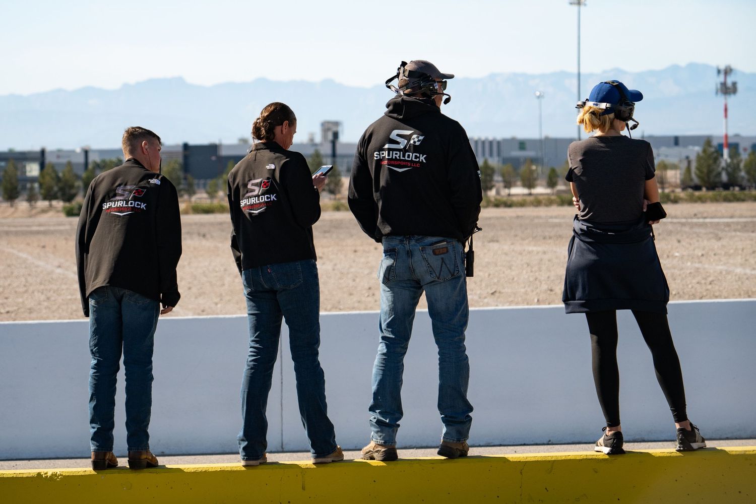 Four people wearing Spurlock Racing jackets watching race cars from pit lane at a desert track.