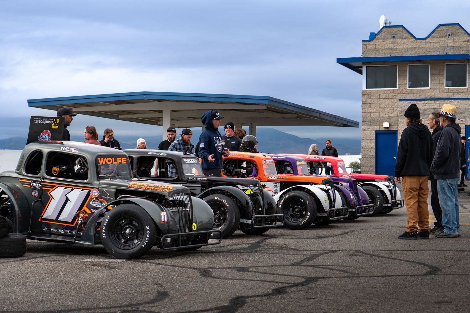 A lineup of colorful vintage-style legend race cars parked at a racing circuit on an overcast day.