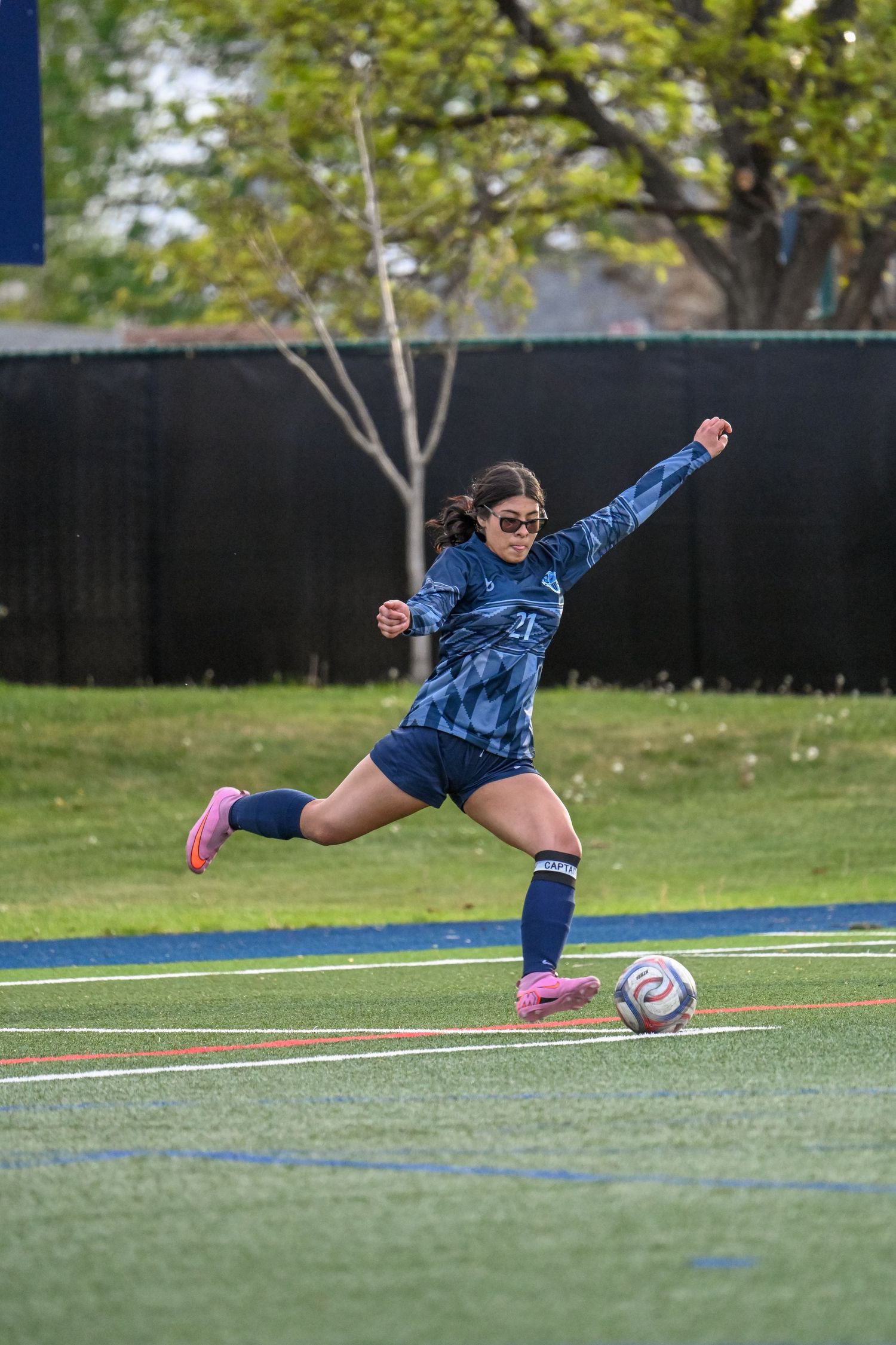 Female soccer player in navy uniform kicking ball on green turf field during outdoor match.