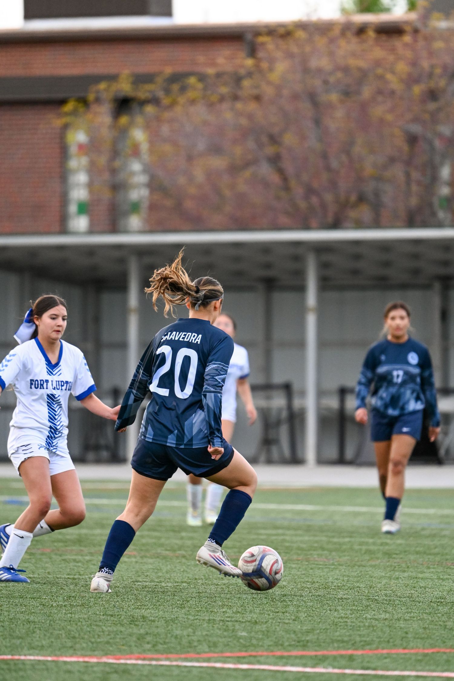 Player #20 Saavedra dribbles soccer ball in women's match, competing against Fort Lewis College defender.